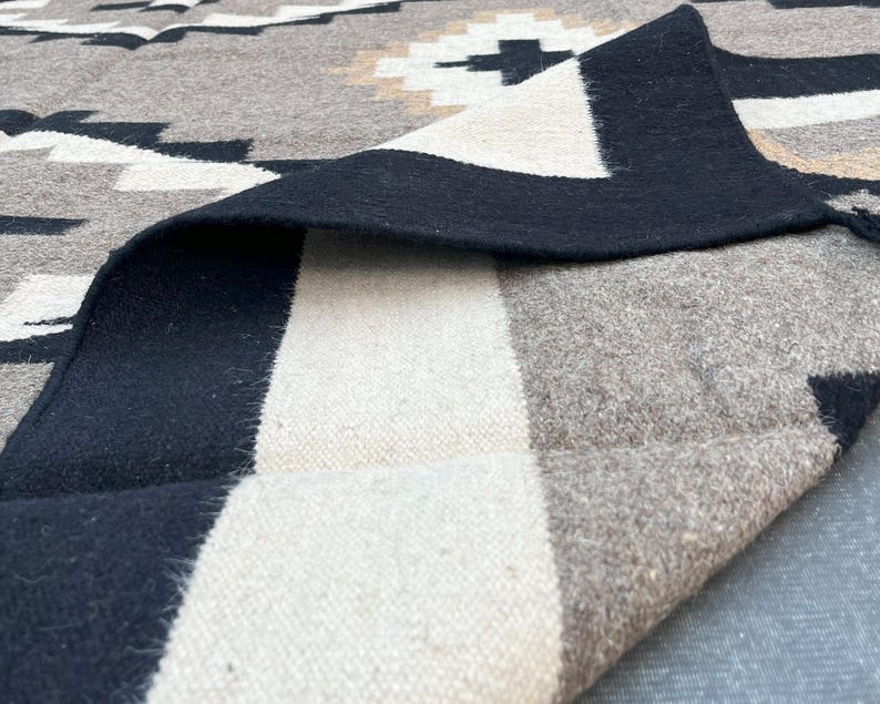 May include: A folded, patterned blanket in shades of gray, black, and cream. The design features geometric shapes and a black border. The blanket's texture is visible, suggesting a woven or knitted material. The overall aesthetic is rustic and cozy.