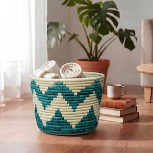 May include: A round woven basket featuring a repeating teal and cream chevron pattern. The basket is filled with several rolled cream-coloured fabric items, showcasing a textured, coiled construction with a wide opening and a flat base.