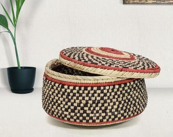 May include: A round, lidded basket featuring a woven checkerboard pattern in black and natural tan fibers. The lid and base are accented with concentric rings of deep red and natural tan, creating a structured, cylindrical storage container.