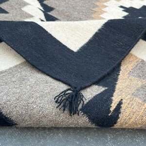 May include: A folded woven blanket with a geometric pattern in black, white, tan, and gray. The blanket has a black border with a fringe of black tassels. The design features triangles and other shapes.