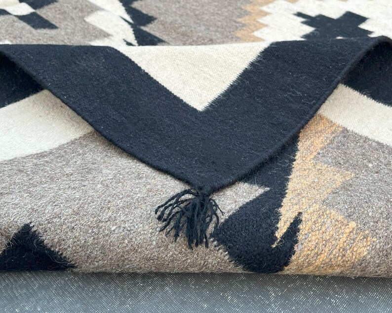 May include: A folded woven blanket with a geometric pattern in black, white, tan, and gray. The blanket has a black border with a fringe of black tassels. The design features triangles and other shapes.