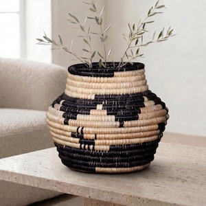 May include: A woven basket vase with a black and natural color geometric pattern. The vase is filled with green leafy branches. The vase sits on a light-colored stone table, with a neutral-toned sofa in the background.