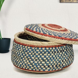 May include: A round, lidded storage basket crafted from woven natural fibers. The basket features a repeating checkered pattern in shades of blue and cream, accented by horizontal bands of terracotta red at the base, rim, and lid center.