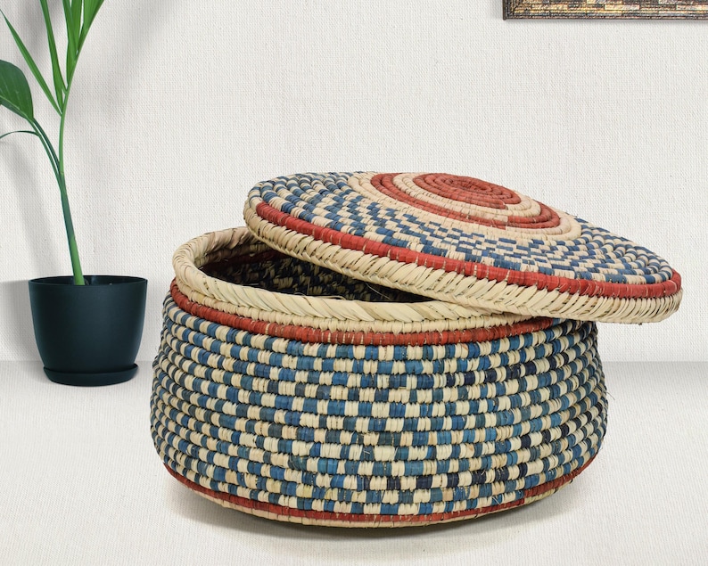 May include: A round, lidded storage basket crafted from woven natural fibers. The basket features a repeating checkered pattern in shades of blue and cream, accented by horizontal bands of terracotta red at the base, rim, and lid center.