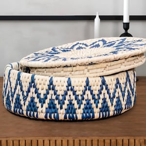 May include: A round woven basket with a lid. The basket and lid are made of natural-colored fibers with a blue geometric pattern. The basket is sitting on a wooden surface.