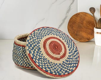 May include: A round woven basket with a lid. The lid and sides of the basket are decorated with a blue and white checkered pattern, with a red spiral design in the center. The basket is open, revealing the interior.