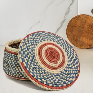 May include: A round woven basket with a matching lid, featuring a concentric pattern of red, blue, and natural beige fibers. The basket has a coiled construction with a flat top and a slightly tapered body, showcasing a detailed, multi-colored design.