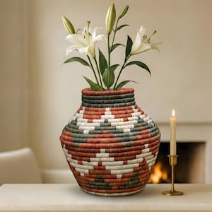 Puede incluir: Un jarrón tejido con un patrón geométrico en rojo, blanco y verde contiene lirios blancos. Una vela encendida en un soporte dorado está cerca. El jarrón está sobre una mesa blanca.