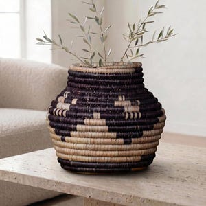 May include: A woven vase with a dark brown and natural color geometric pattern. The vase is filled with green leafy branches. The vase is sitting on a light-colored stone table. Home decor.