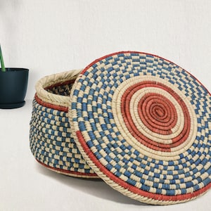 May include: A round, lidded basket crafted from coiled natural fibers, featuring a geometric pattern in red, blue, and cream. The lid displays concentric circles of red at the center, surrounded by alternating blue and cream segments, while the basket base mirrors this woven color scheme.