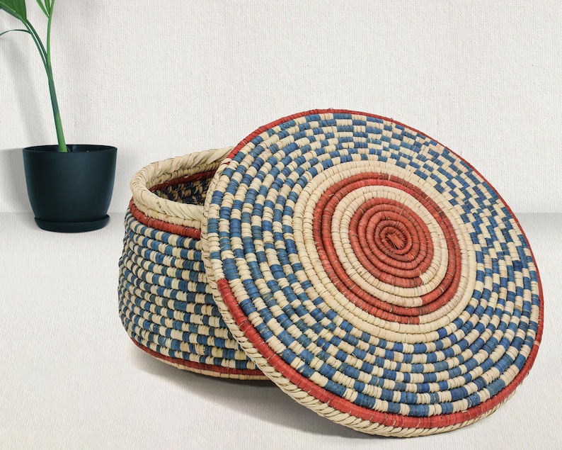 May include: A round, lidded basket crafted from coiled natural fibers, featuring a geometric pattern in red, blue, and cream. The lid displays concentric circles of red at the center, surrounded by alternating blue and cream segments, while the basket base mirrors this woven color scheme.