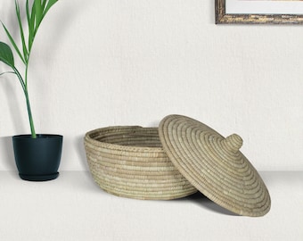 May include: A round, shallow, light tan woven basket with a matching conical lid featuring a small central knob. The basket has a coiled texture and a flat base, constructed from natural dried plant fibers, presented as a storage container.