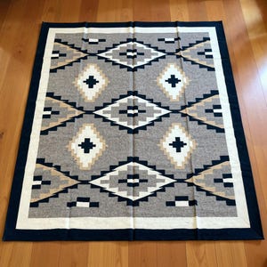 May include: A rectangular gray blanket with a geometric pattern. The design features diamond shapes in white, black, and tan, with a navy blue border and white inner border. The blanket is folded neatly on a wooden floor.
