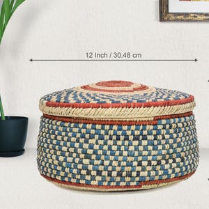 May include: A round woven basket with a lid, featuring a checkered pattern in blue, cream, and red. The basket measures 12 inches in diameter and 8 inches in height. The lid has a circular red center, and the base is a flat, circular shape.