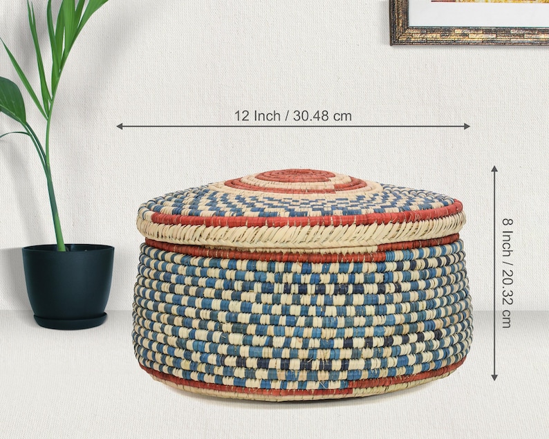 May include: A round woven basket with a lid, featuring a checkered pattern in blue, cream, and red. The basket measures 12 inches in diameter and 8 inches in height. The lid has a circular red center, and the base is a flat, circular shape.