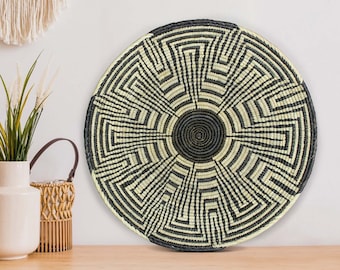 May include: A round, woven wall basket with a geometric design. The basket features a central spiral in dark gray, surrounded by concentric patterns in alternating black and cream colors. The basket is a decorative item.