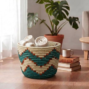 May include: A round woven basket featuring a geometric pattern in teal and terracotta on a cream-coloured base. The basket is filled with several rolled-up cream-coloured fabric items, showcasing a textured, coiled construction throughout the exterior.
