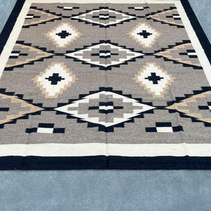 May include: A rectangular rug with a geometric pattern. The rug features a central design of diamonds and crosses in shades of gray, black, white, and tan. A black border frames the design, with a white and tan border inside.