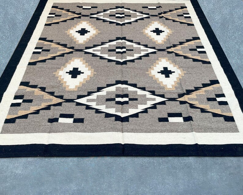 May include: A rectangular rug with a geometric pattern. The rug features a central design of diamonds and crosses in shades of gray, black, white, and tan. A black border frames the design, with a white and tan border inside.