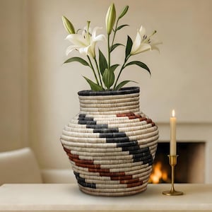 May include: A woven basket vase with a black, white, and red geometric pattern, holding white lilies. A lit candle in a gold-colored holder sits nearby. The vase is on a white surface.