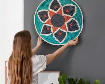 May include: A round, woven wall basket with a turquoise border and a central floral design in orange, navy, and white. The basket is being held up against a gray wall, showcasing its decorative pattern and texture.