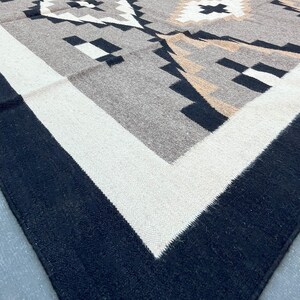 May include: A woven rug with a geometric pattern in shades of gray, black, and beige. The rug has a black border, a white inner border, and a central design featuring diamond and arrow shapes.