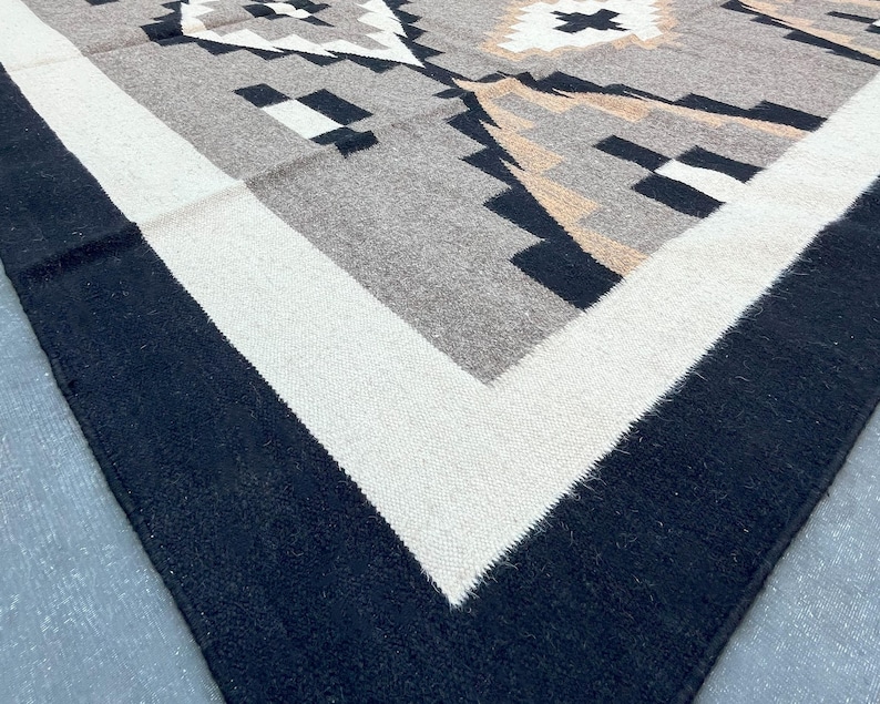 May include: A woven rug with a geometric pattern in shades of gray, black, and beige. The rug has a black border, a white inner border, and a central design featuring diamond and arrow shapes.