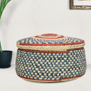 May include: A round, lidded storage basket featuring a woven pattern in alternating blue, cream, and red fibers. The basket has a flat, circular lid with a concentric red center, and the sides display a consistent checkered weave design.