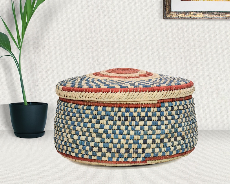May include: A round, lidded storage basket featuring a woven pattern in alternating blue, cream, and red fibers. The basket has a flat, circular lid with a concentric red center, and the sides display a consistent checkered weave design.