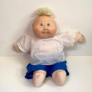 Puede incluir: Una muñeca vintage con la cabeza calva y un mechón de pelo amarillo claro. La muñeca tiene ojos azules y una expresión sonriente. Lleva una camisa blanca y pantalones cortos azules. Los brazos de la muñeca están extendidos y se ven sus pies.