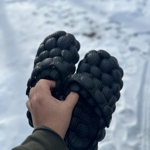 Peut inclure: Une paire de tongs à bulles noires avec un design texturé. Les tongs sont tenues dans une main.