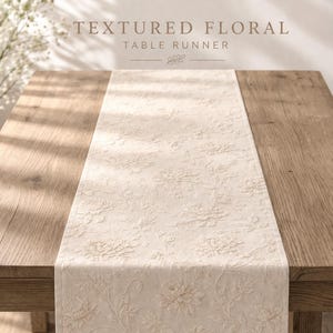 Puede incluir: Camino de mesa floral texturizado de color crema sobre una mesa de madera. El camino de mesa presenta un patrón repetido de flores y follaje en relieve. El texto "TEXTURED FLORAL TABLE RUNNER" está en la parte superior de la imagen.