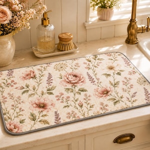 Puede incluir: Un tapete rectangular para secar la cocina con un estampado floral de rosas rosas y ramitas de lavanda sobre un fondo crema. El tapete tiene un borde gris y está colocado sobre una encimera blanca cerca de un fregadero.