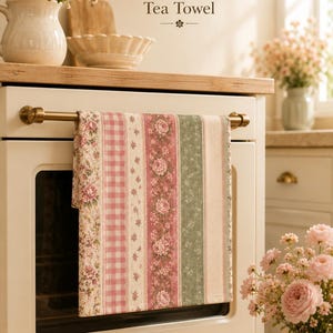 May include: A cotton tea towel with a floral and striped pattern in pink, green, and cream hangs on a brass bar. The towel is displayed in a kitchen setting with flowers and a white oven.