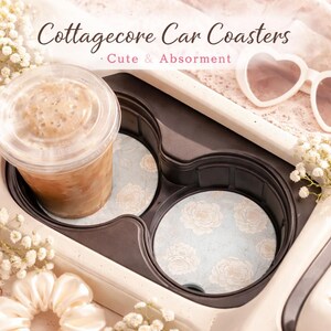May include: Cottagecore car coasters with a floral design. The coasters are light blue with a pattern of white flowers. A cup of iced coffee sits in the car's cup holder. The text on the image says "Cottagecore Car Coasters - Cute & Absorbent."