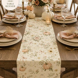 Puede incluir: Un camino de mesa color crema de 40,64 cm por 182,88 cm, con un patrón repetitivo de pequeños fantasmas, calabazas rosa claro, ramas de flores y lazos. La tela está extendida sobre una mesa de comedor de madera con platos y vasos.