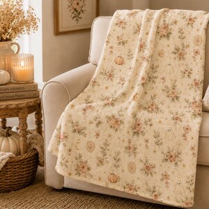 Decorative floral ghost blanket styled on a cream sofa with blush pumpkins, candles, woven textures, and soft seasonal home accents.