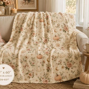 Summerween plush blanket draped across a cream sofa featuring blush florals, ghosts, pumpkins, bows, and moon details in a cozy cottage-style living room.