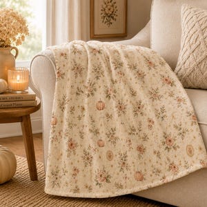 Summerween throw blanket draped over a cream sofa arm beside candles, books, and neutral autumn decor in a cozy cottage living room.