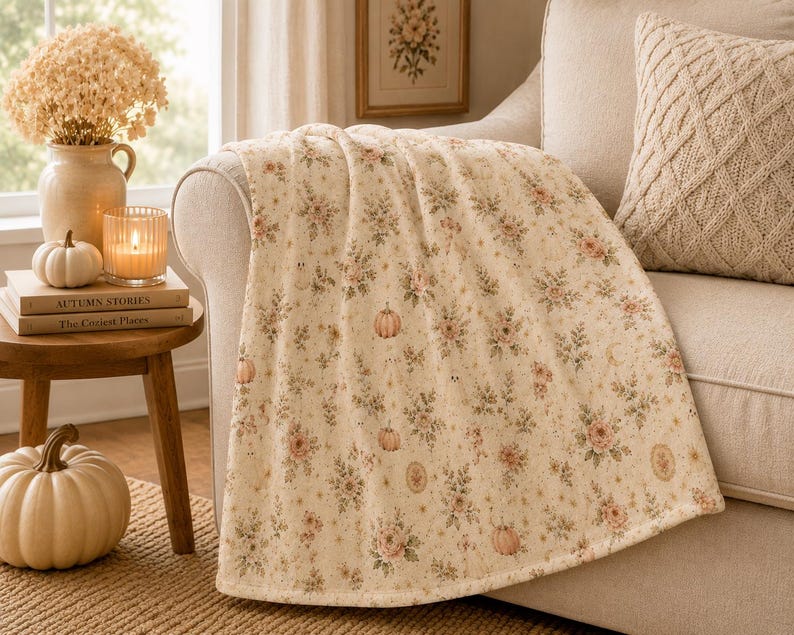 Summerween throw blanket draped over a cream sofa arm beside candles, books, and neutral autumn decor in a cozy cottage living room.