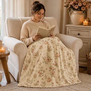 Woman reading in a cream armchair with a Summerween floral ghost blanket draped across her lap in a warm neutral cottage-inspired space.
