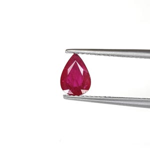 May include: A pear-shaped, faceted ruby gemstone with a deep red color held by a pair of silver-toned metal tweezers. The gemstone features a pointed top and a rounded base, displaying multiple geometric facets that reflect light across its surface.