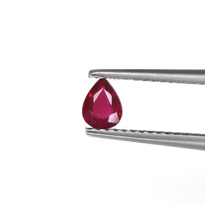 Può includere: Una gemma rosso intenso a forma di pera con superficie sfaccettata, tenuta da una pinzetta in metallo color argento. La pietra presenta una qualità traslucida con tagli geometrici netti che riflettono la luce sulla sua superficie lucida.