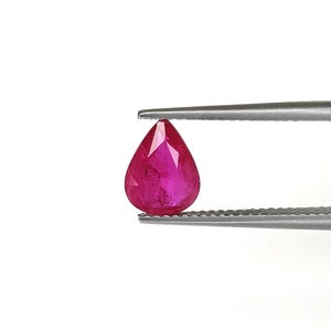 May include: A pear-shaped, faceted ruby gemstone with a vibrant, deep pinkish-red color. The gemstone is held between the serrated metal tips of a pair of silver-toned tweezers, showcasing its clear, polished surface and precise, symmetrical cut.