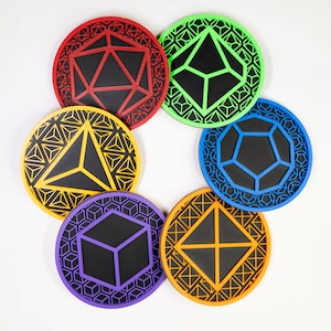 May include: A collection of seven colorful, round coasters with geometric designs. Each coaster features a different shape, including a cube, pyramid, and a 20-sided die. The coasters are in red, green, yellow, blue, orange, and purple, with black centers.