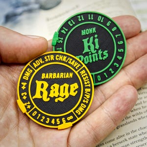 May include: Two circular game tokens are held in a hand. One is black with yellow text reading "Barbarian Rage". The other is black with green text reading "Monk Ki Points". Both have numerical markings around the edge.