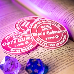 May include: Four pink and white round game tokens with text that reads "Gain Adv. On Con Checks Enhance Ability: Bear's Endurance Gain 2D6 Temp HP When Cast 1 Hour" and "Gain Adv. On Con Checks Enhance Ability: Owl's Wisdom For The Duration 1 Hour".
