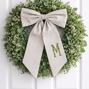 Op de afbeelding: Een decoratieve krans met weelderig groen en kleine witte bloemen. Een grote, gestreepte strik met een groen geborduurde "M" voegt een persoonlijk tintje toe. De krans hangt aan een witte deur.