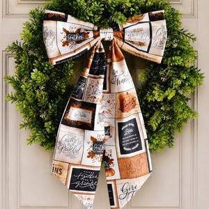 May include: A circular green boxwood wreath adorned with a large fabric bow featuring a grid pattern of autumn-themed text and illustrations including the words Gather, Grateful, Blessed, Thankful, and Give, displayed on a paneled door.