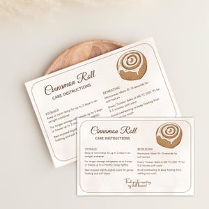 May include: Two cream-colored cards with the text "Cinnamon Roll Care Instructions" and an illustration of a cinnamon roll. The cards provide storage and reheating instructions. The cards are on a wooden plate.
